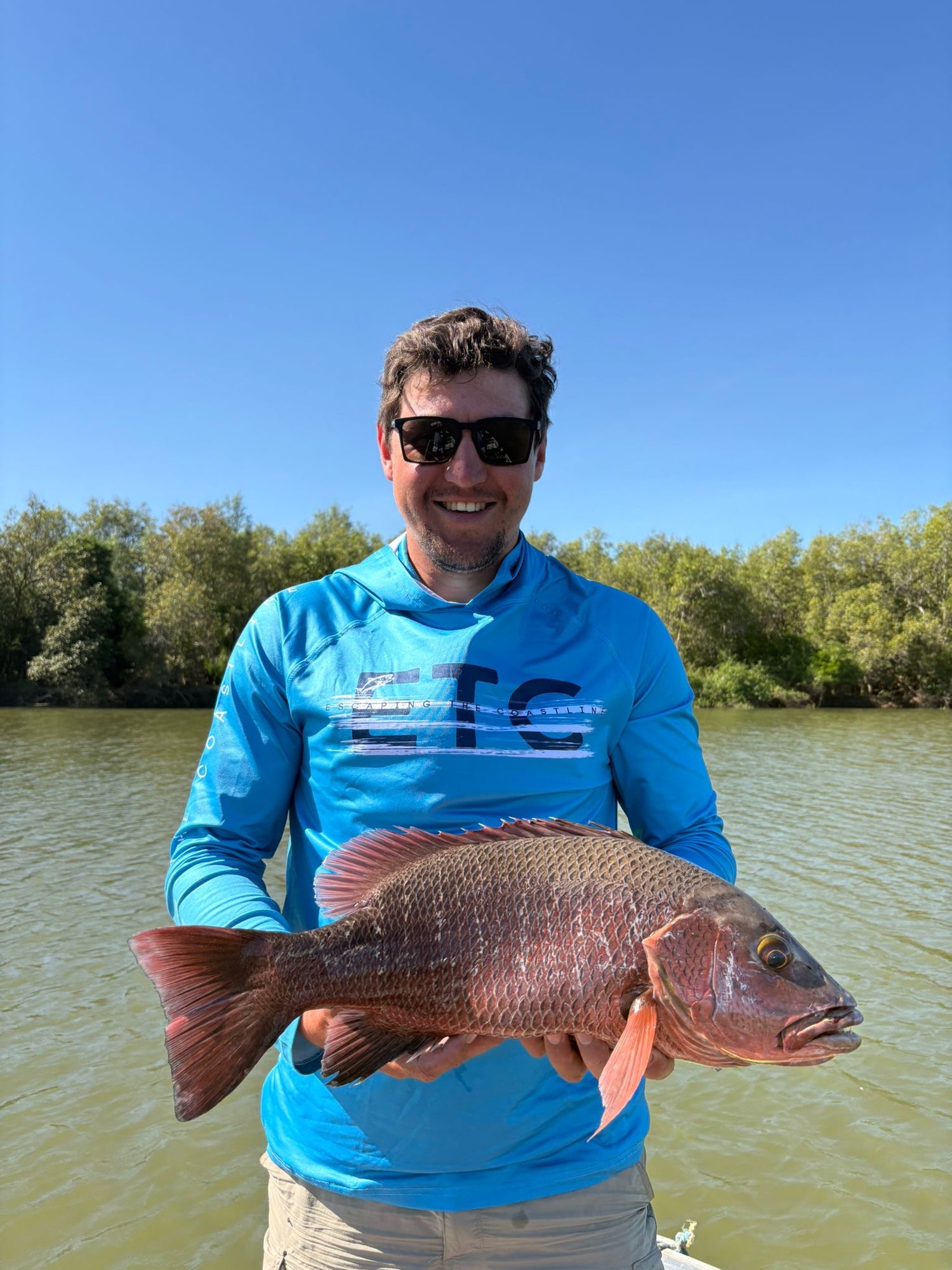 Man holding a large Mangrove Jack by a body of water with trees in the background wearing a blue Escaping The Coastline (ETC) fishing shirt with hoof & Buff