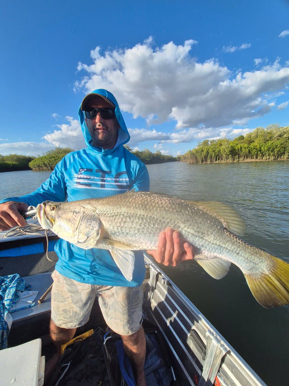 Man holding a large barramundi on a boat with a scenic background wearing a blue Escaping The Coastline (ETC) Tide Chaser Performance fishing shirt with hood & Buff