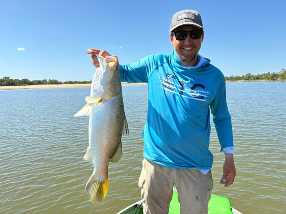 Man holding a Barramundi on a lake with a clear sky, wearing a blue Escaping The Coastline (ETC) Tide Chaser high performance fishing shirt with hood & Buff