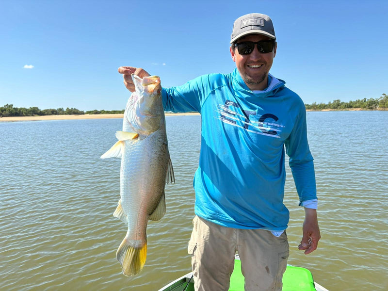Man holding a Barramundi on a lake with a clear sky, wearing a blue Escaping The Coastline (ETC) Tide Chaser high performance fishing shirt with hood & Buff