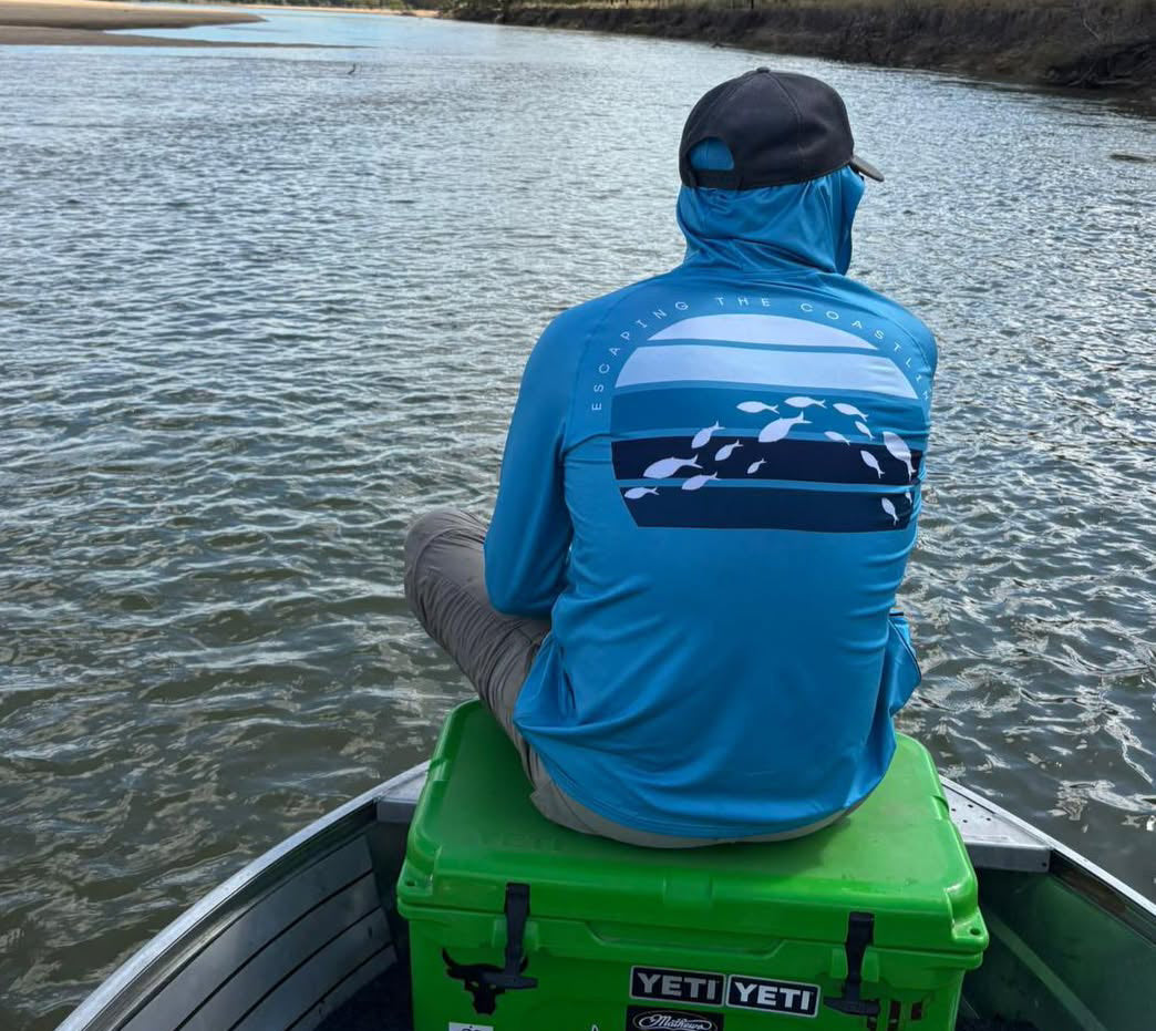 Person wearing a blue Escaping The Coastline (ETC) Tode Chaser Preformance Fishing Shirt with Hood & Buff, sitting on a green YETI cooler by a body of water.