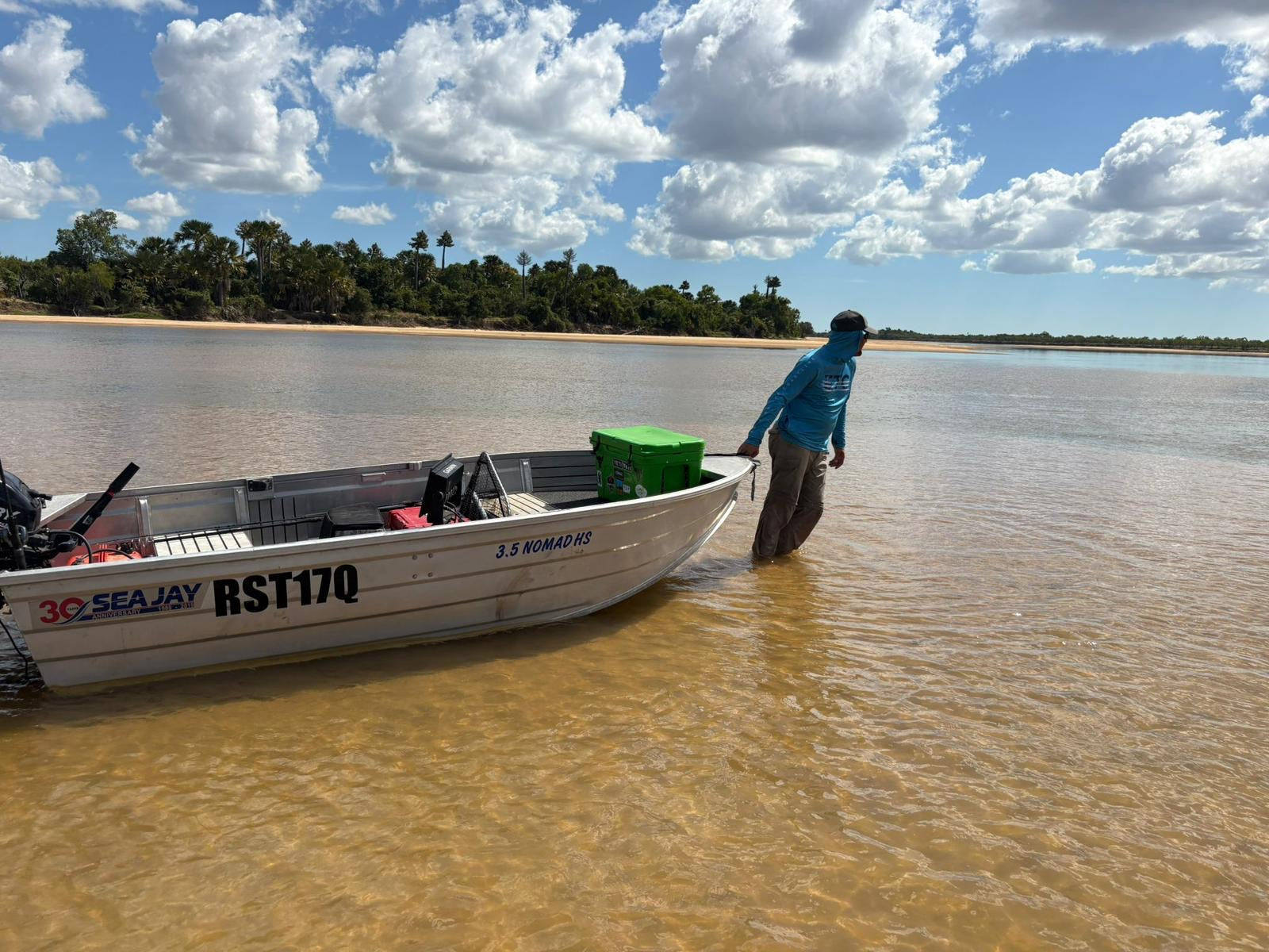 Person standing next to a boat on a body of water with Escaping The Coastline Shirt with trees and sky in the background