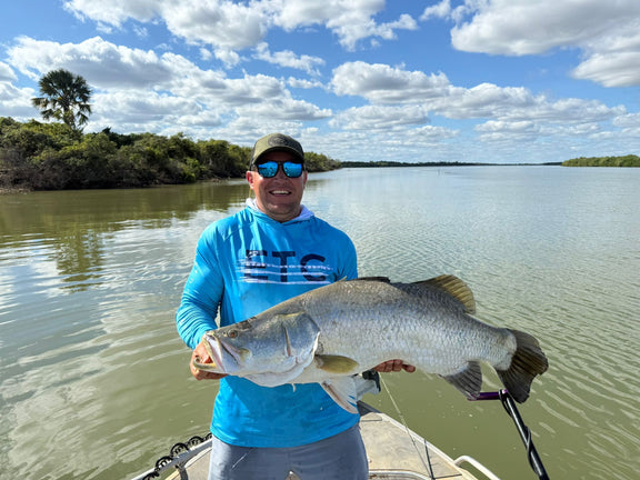Man holding a large Barramundi fish on a boat with a scenic lake background wearing a blue Escaping The Coastline (ETC) fishing Shirt with hood and buff