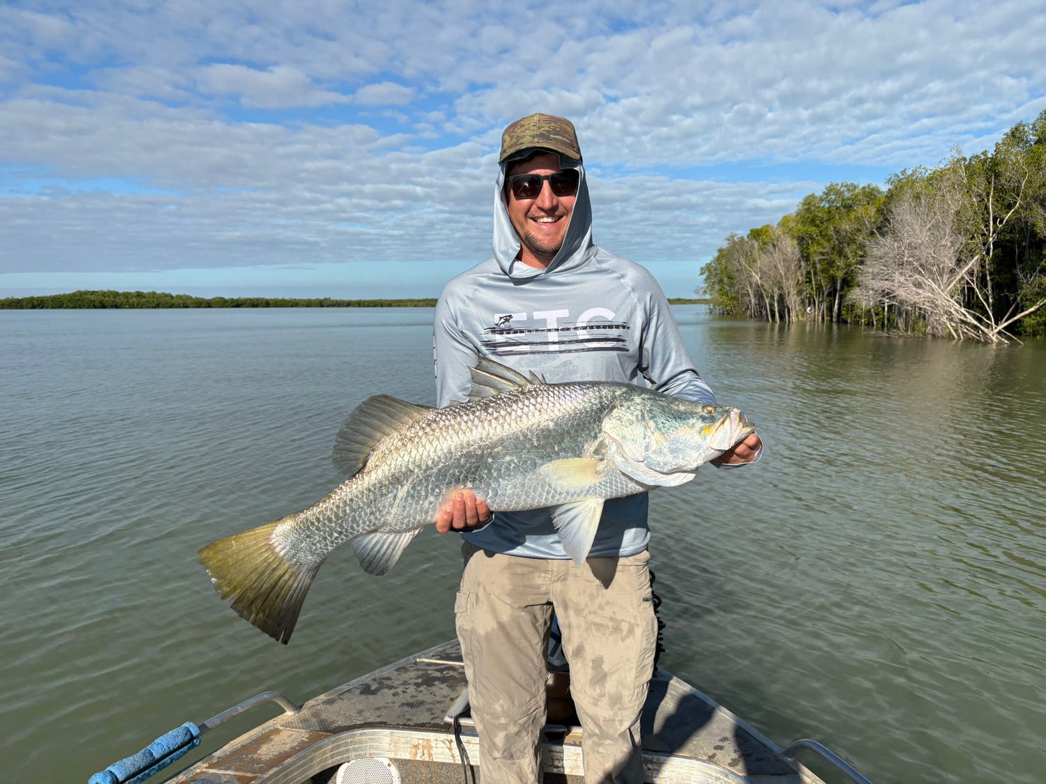Man holding a Barramundi large fish on a boat with a scenic background in far north queensland, Australia wearing a grey Escaping The Coastline (ETC) reef reaper buff and hooded fishing shirt