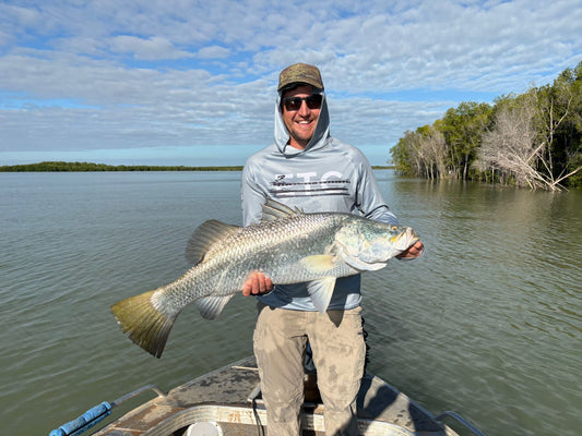 Man holding a Barramundi large fish on a boat with a scenic background in far north queensland, Australia wearing a grey Escaping The Coastline (ETC) reef reaper buff and hooded fishing shirt
