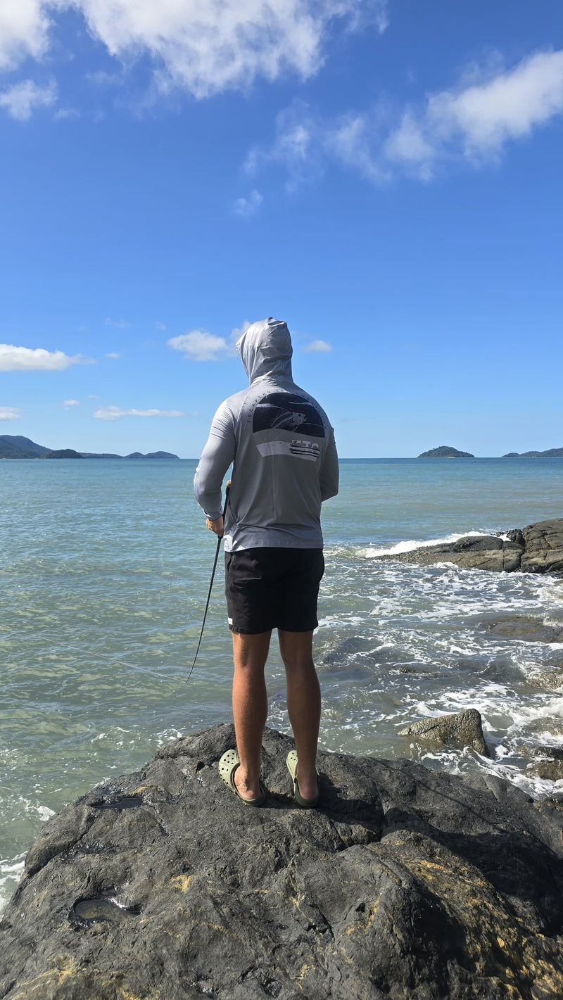 Person standing on a rock by the ocean with a clear blue sky. Cathching fish wearing a grey Escaping The Coastline (ETC) shirt with a buff and hood.