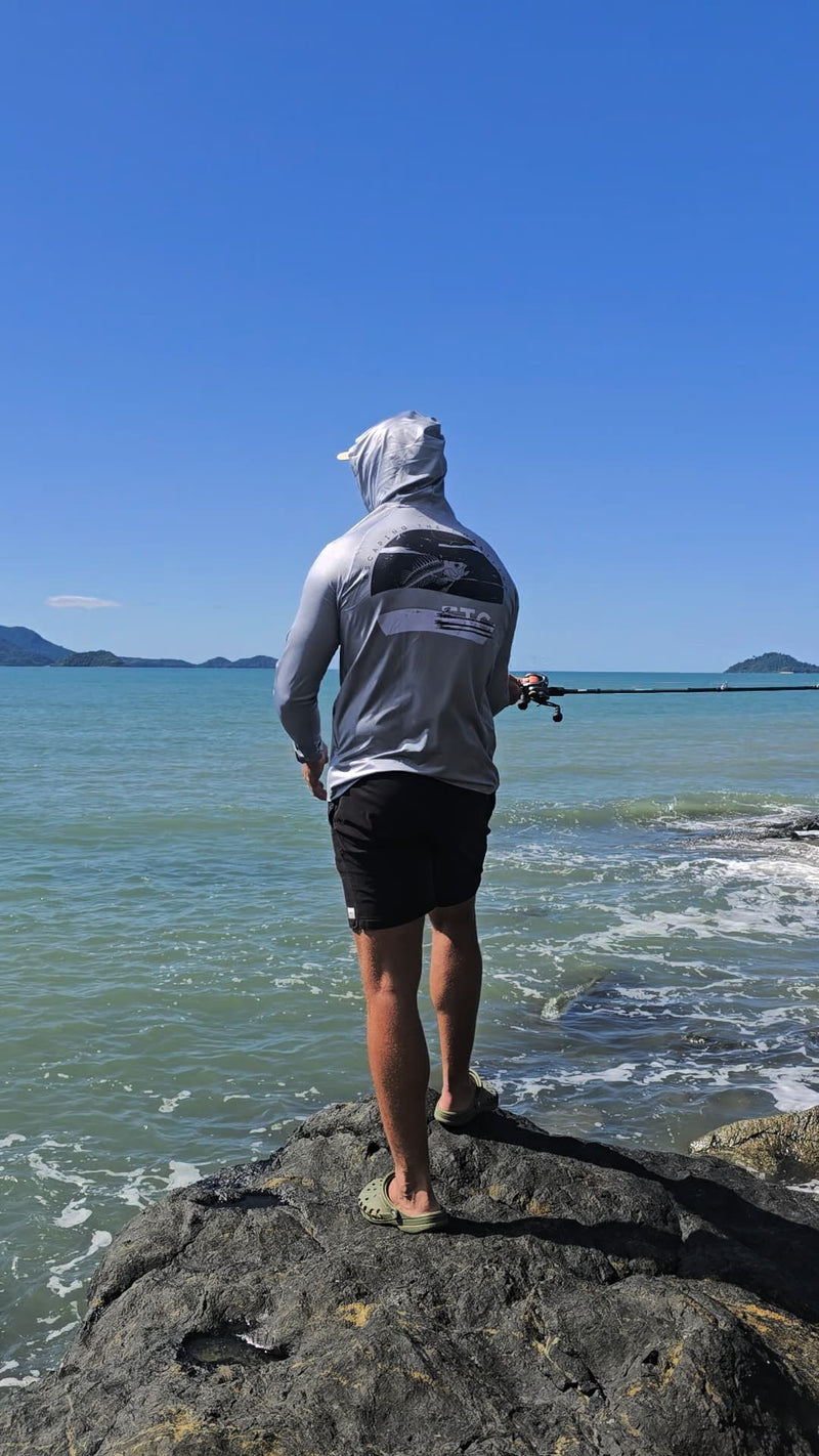 Person fishing from a rock on a beach with clear blue sky wearing Escaping The Coastline (ETC) grey Reef Reaper Buff and Hooded shirt