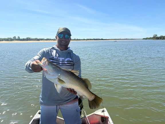 Man holding a barramundi on a boat with a lake in the background, wearing a Grey Escaping The Coastline (ETC) shirt with buff & Hood