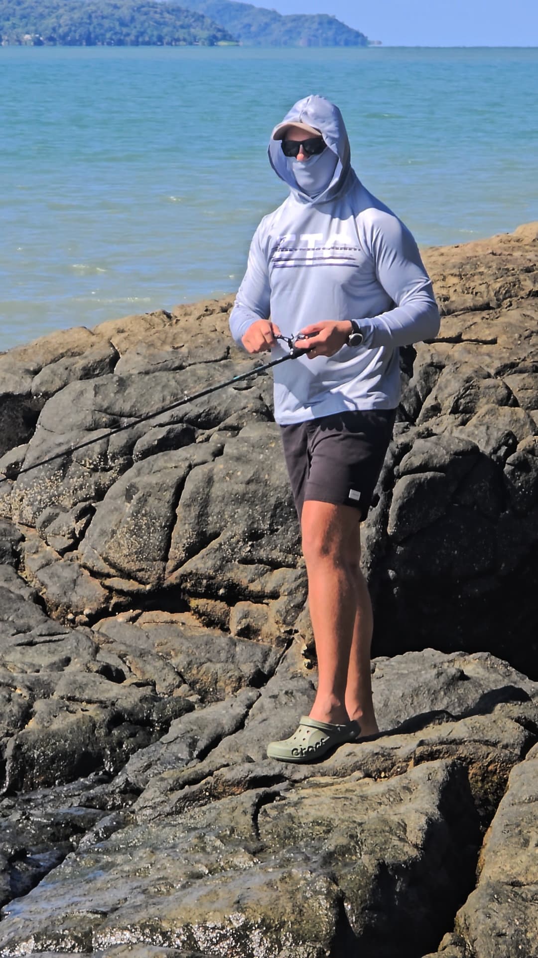 Person fishing on rocky coastal area with ocean and sky in the background, wearing a grey reef reaper Escaping The Coastline (ETC) fishing Shirt with buff & hood