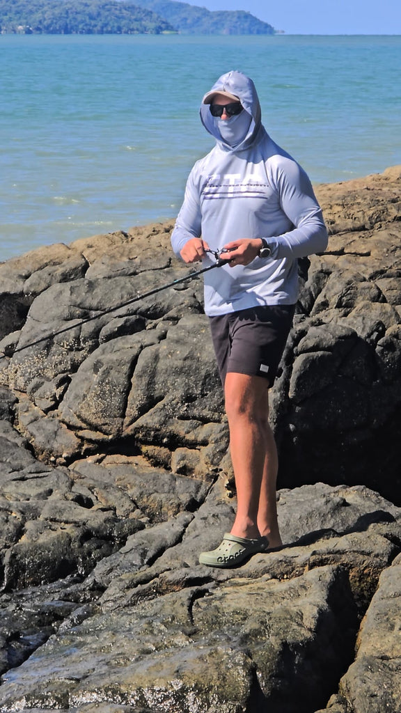 Person fishing on rocky coastal area with ocean and sky in the background, wearing a grey reef reaper Escaping The Coastline (ETC) fishing Shirt with buff & hood